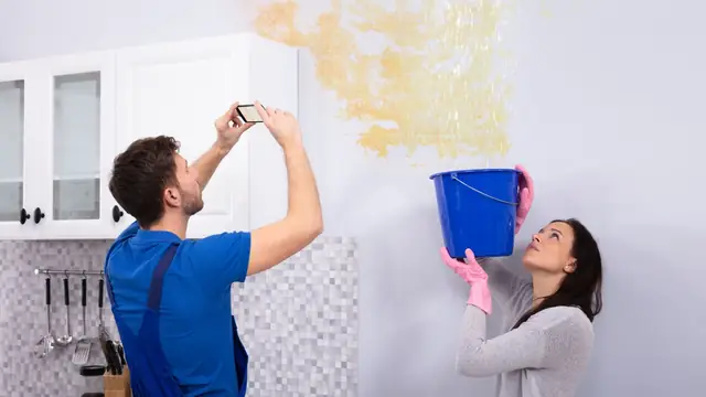 Man photographing a water-stained wall and ceiling with his phone while a woman holds a bucket below
