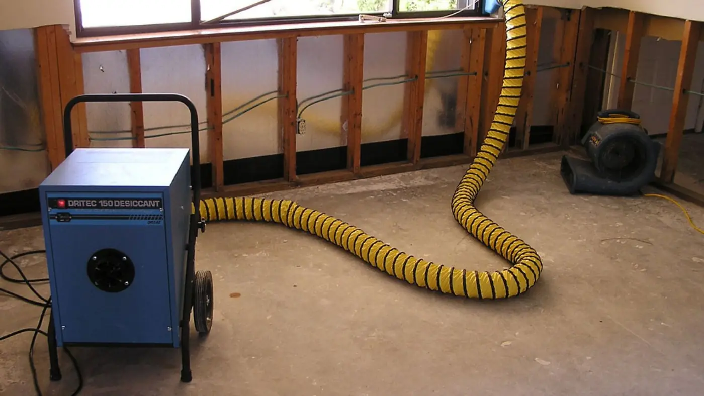Dehumidifier with a yellow duct hose and an air mover in a room where the lower wall has been opened to dry